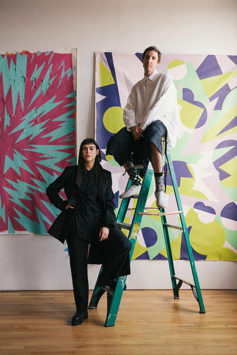 Two people pose in front of abstract colorful paintings; one stands on the floor, the other sits on a green ladder. The setting appears to be an art studio.