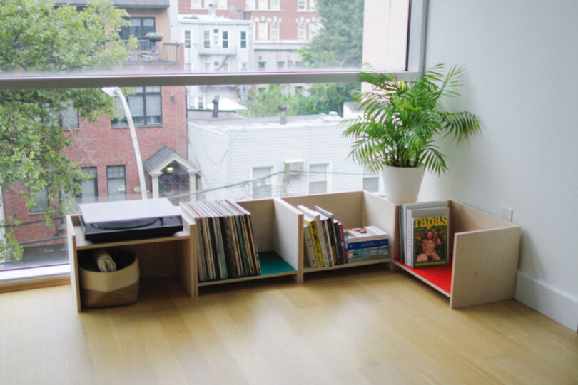 Low wooden shelves by a large window from Elements Studio hold a record player, vinyl records, books, a basket, a potted plant, and a magazine in a bright, modern room with hardwood floors