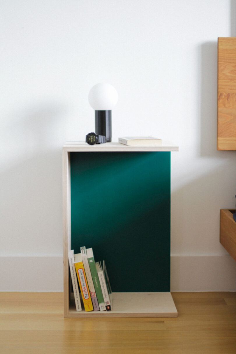 A minimalist nightstand holds books, a black clock, a closed book, and a small lamp, set beside a wooden bed against a white wall.