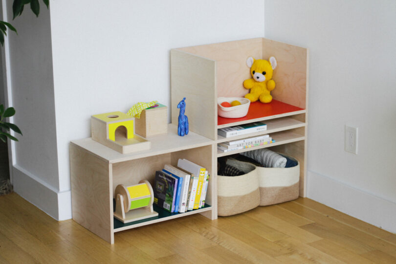 A wooden shelf unit holds a yellow stuffed toy, books, baskets, and small toys, positioned in the corner of a room with light wood flooring