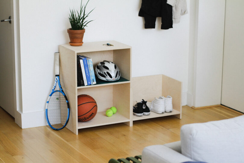 In this tidy, modern room, a wooden shelf holds books, a plant, a bicycle helmet, a basketball, and tennis balls. Rackets, shoes, and jackets are neatly arranged throughout the space