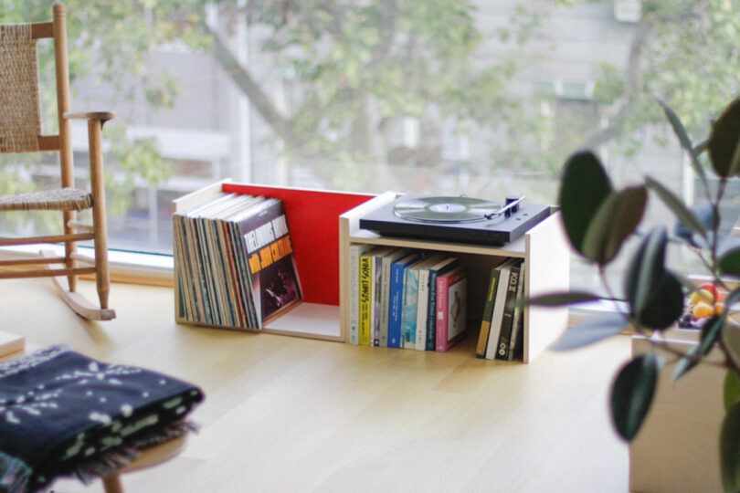 A record player from Elements Studio sits on a modern shelf holding vinyl records and books next to a large window with trees outside.