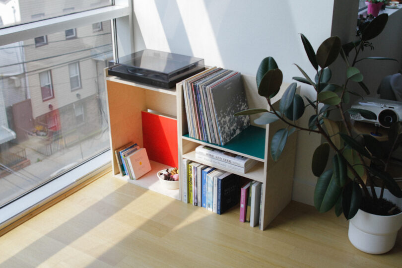 A wooden shelf with a record player, vinyl records, books, and a potted plant beside a large window in a sunlit room