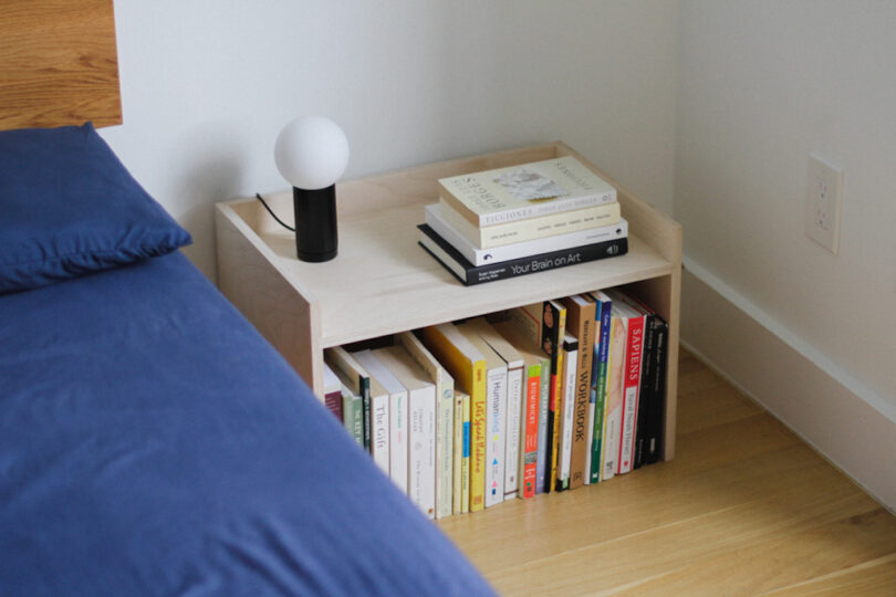 A wooden nightstand sits beside a bed with a blue bedsheet, topped with a round lamp and stacked books, while more books are neatly arranged on the lower shelf.