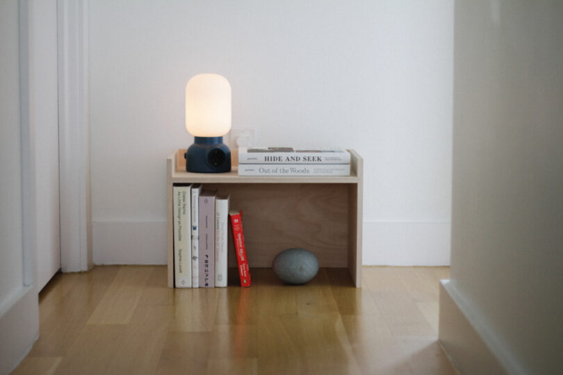 Small wooden bedside table with a lit lamp, several stacked books, a round stone, and a glass of water, placed against a white wall on a wooden floor.