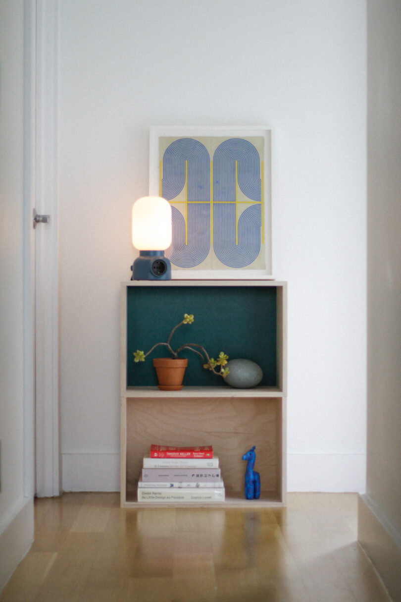 A small wooden shelf holds books, a potted plant, a decorative stone, a blue figurine, and a lit lamp; abstract art is propped on top, against a white wall and wooden floor.