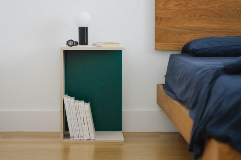 A bedside table with a lamp, a book, and a watch on top, plus several books stored vertically below. It sits next to a wooden bed with dark blue bedding.