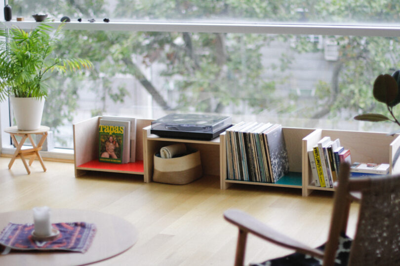 Low wooden shelves with books, magazines, a record player, and a potted plant sit by a large window with trees outside. A small table and chair are in the foreground.