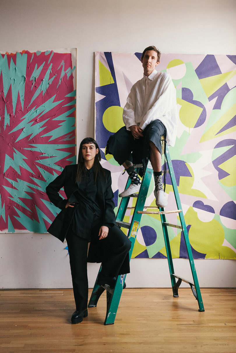 Two people pose in front of abstract colorful paintings; one stands on the floor, the other sits on a green ladder. The setting appears to be an art studio.