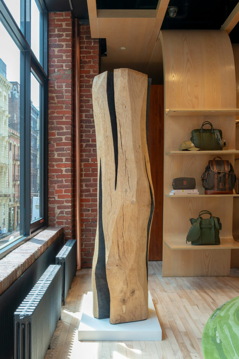 A tall, vertically standing wooden sculpture with organic curves is displayed in a modern store interior near a window and shelves with bags.