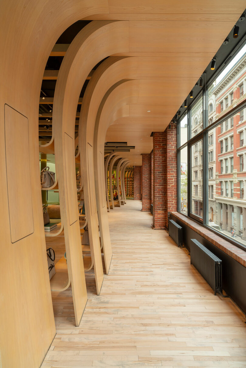 A modern interior hallway with curved wooden arches, large windows, exposed brick walls, and light wood flooring overlooking a city street.