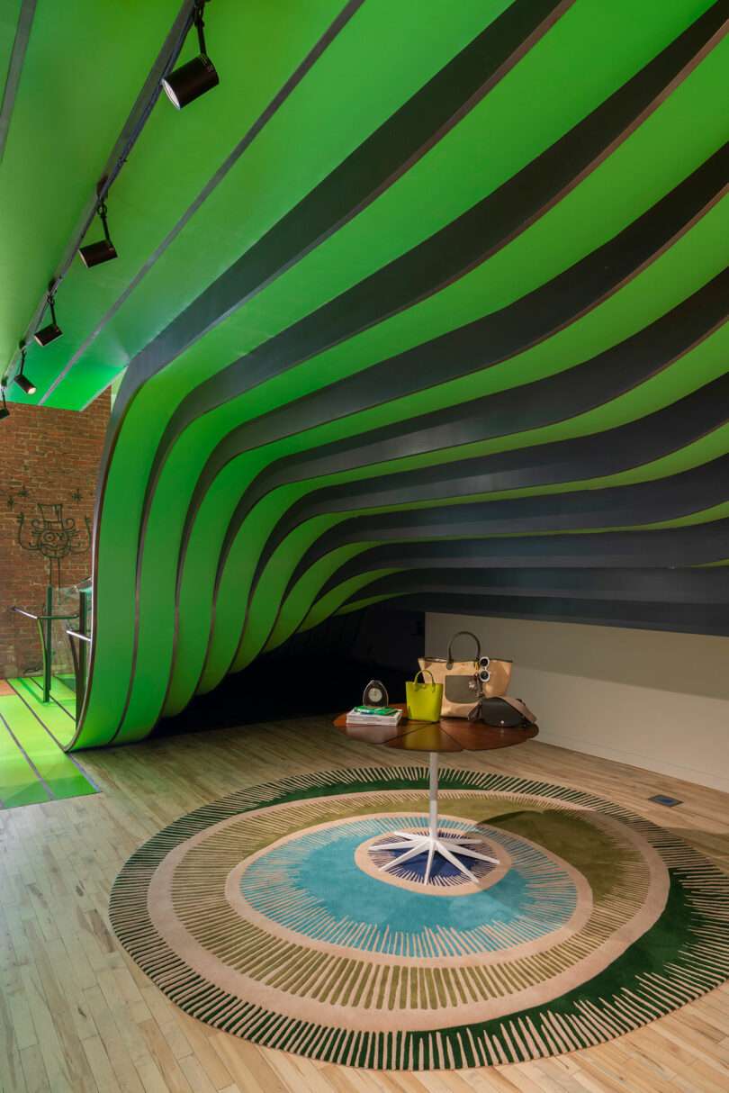 A modern interior with wavy green ceiling panels, a round patterned rug, a small round table displaying handbags, and wooden flooring.