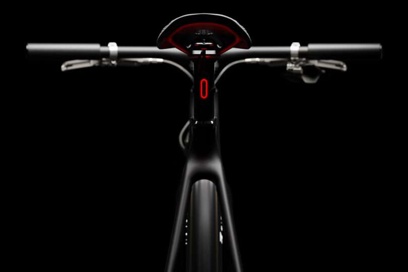 Close-up of a black bicycle viewed from the front, featuring handlebars, seat, and a red digital display on the frame against a black background.