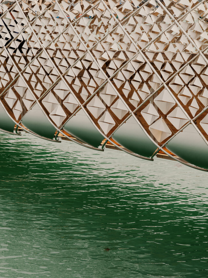 Geometric metal structure with triangular patterns extends over green water, creating reflections on the surface.