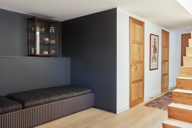 A modern hallway with wooden doors, a built-in bench, a wall-mounted display cabinet, a colorful rug, and a wooden staircase.