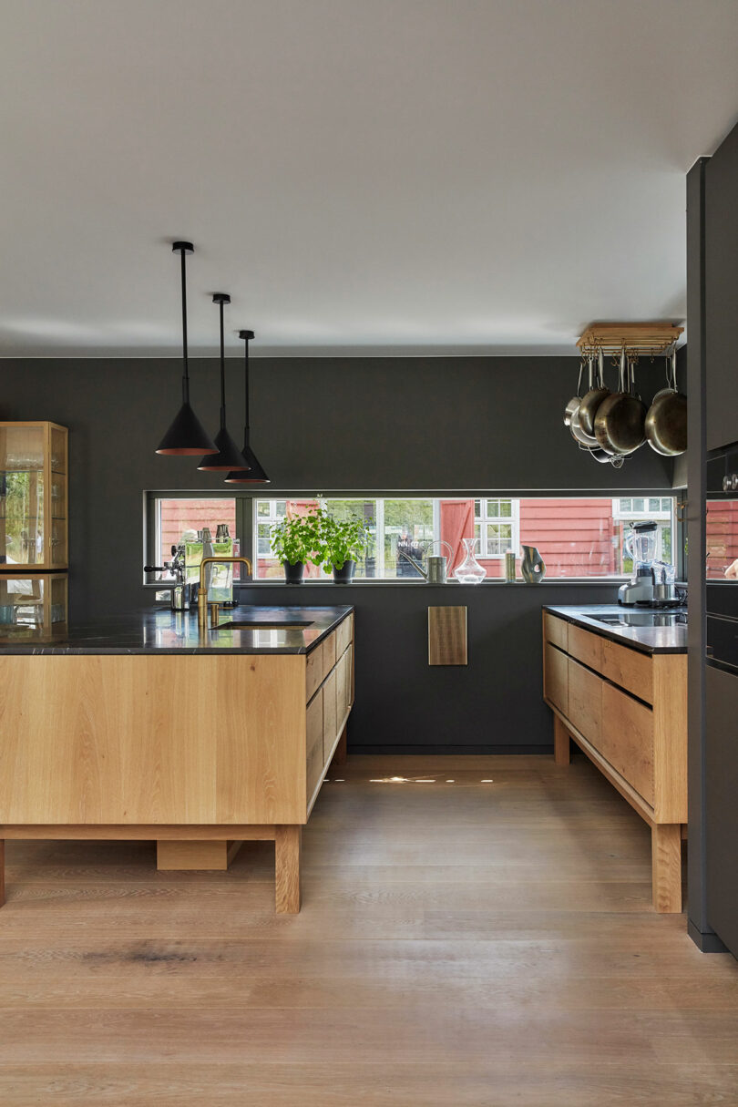 Modern kitchen with wooden cabinets, black countertops, pendant lights, hanging pots, and a large window overlooking a red house and greenery outside.