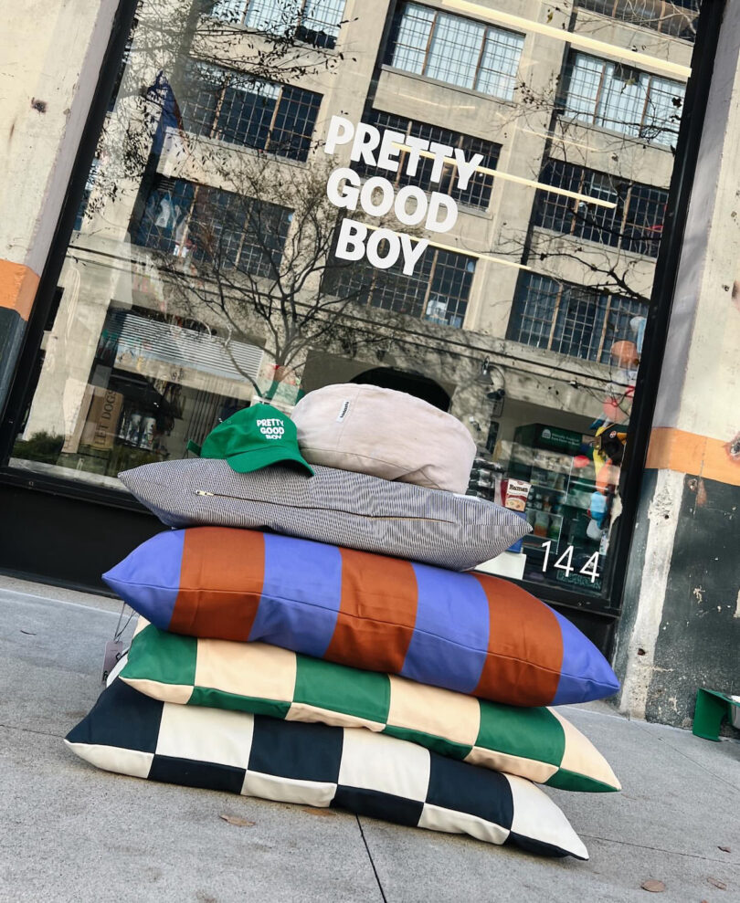 A stack of four striped and checkered cushions, two hats on top, placed on the sidewalk in front of a glass door with 