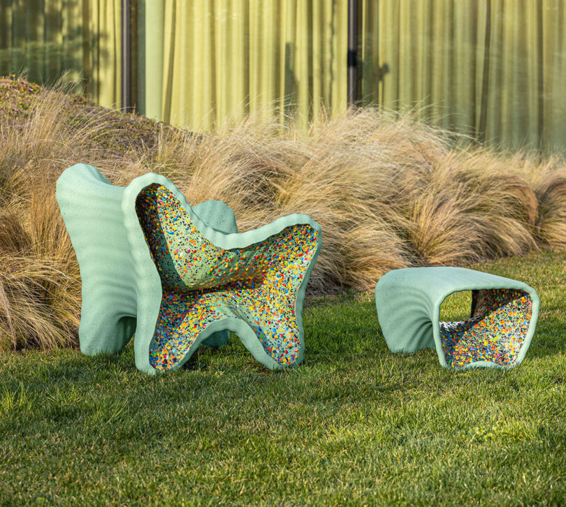 Two sculptural, pastel-green In-Side Thomas Heatherwick outdoor chairs with speckled multicolor interiors and an abstract shape sit on grass in front of tall ornamental grasses and glass windows.