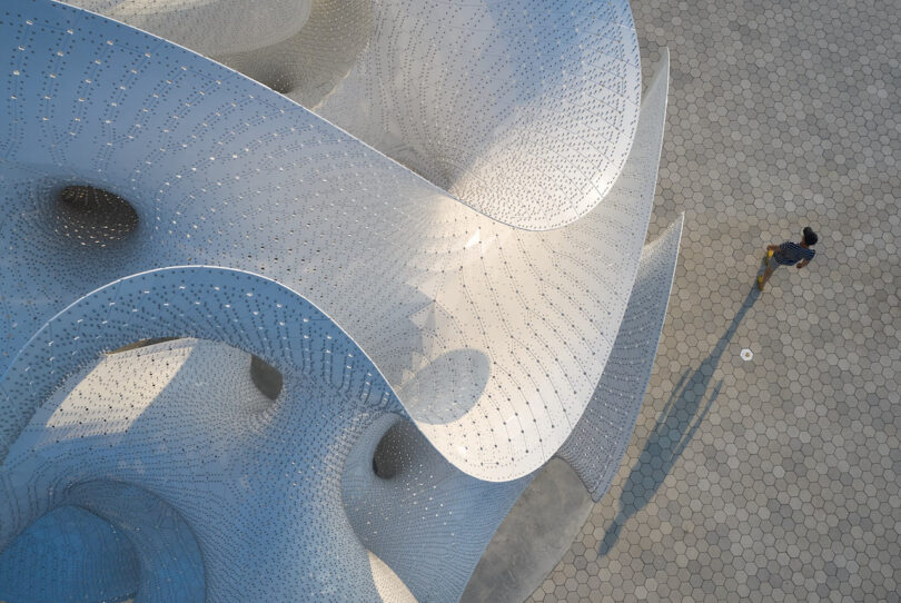A person walks on a patterned pavement next to a large, curved, perforated metal sculpture viewed from above