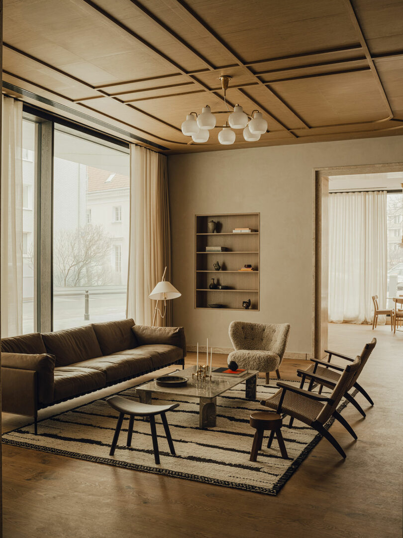 A modern living room with a brown leather sofa, a marble coffee table, armchairs, a striped rug, and large windows with sheer curtains.