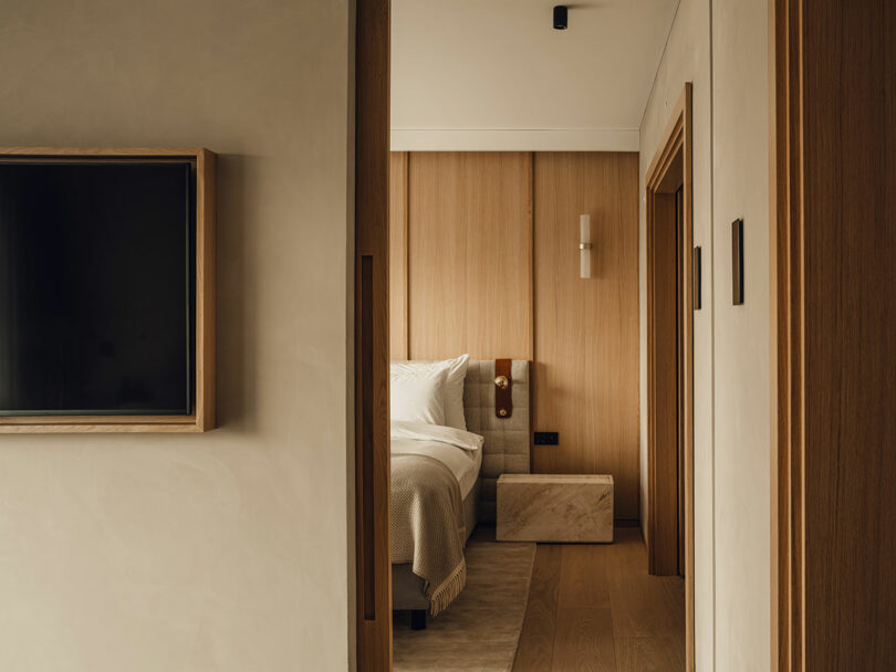 View into a minimalistic bedroom with light wood walls, a neatly made bed, and a mounted TV in the hallway.