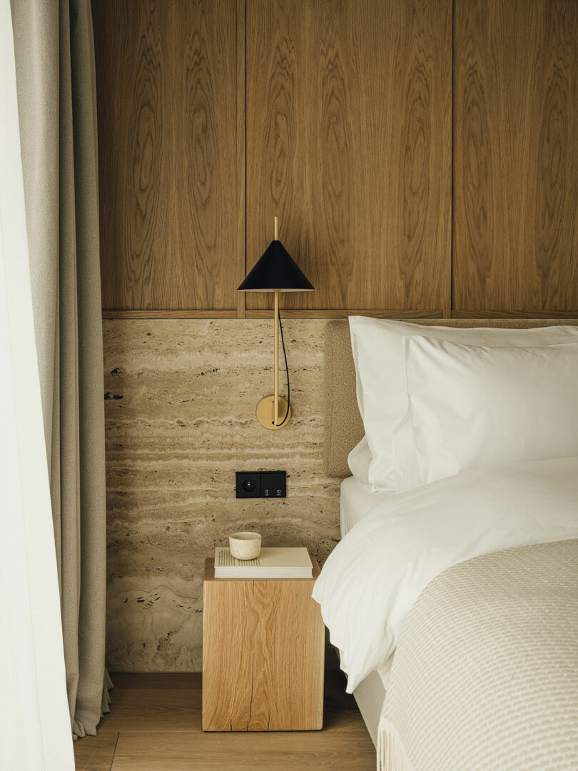 A modern bedroom corner with a wooden headboard, beige bedding, a small wooden side table, a cup, and a black wall-mounted lamp.
