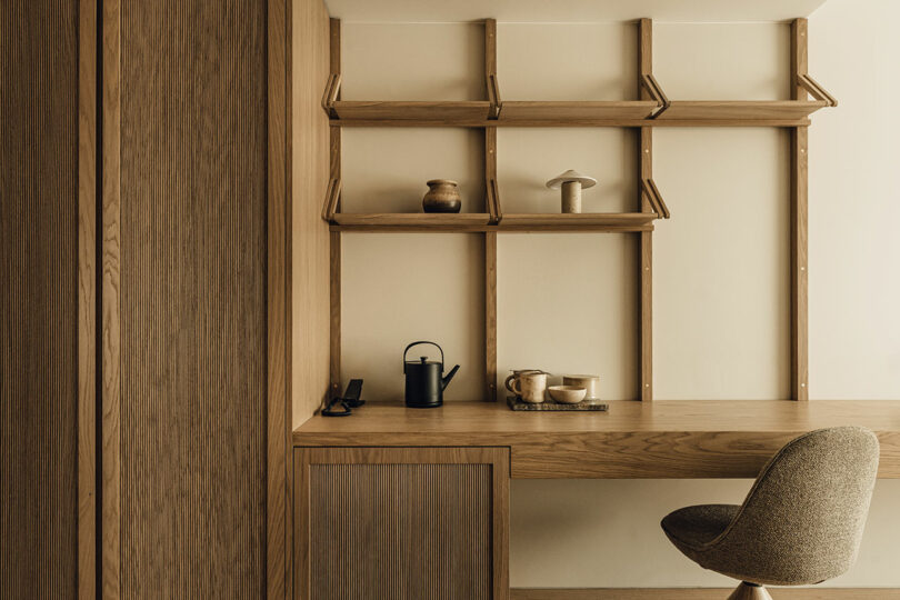 Minimalist wooden desk with built-in shelves, a modern chair, and simple decor items including a teapot, mug, and small vases in a neutral-toned room.