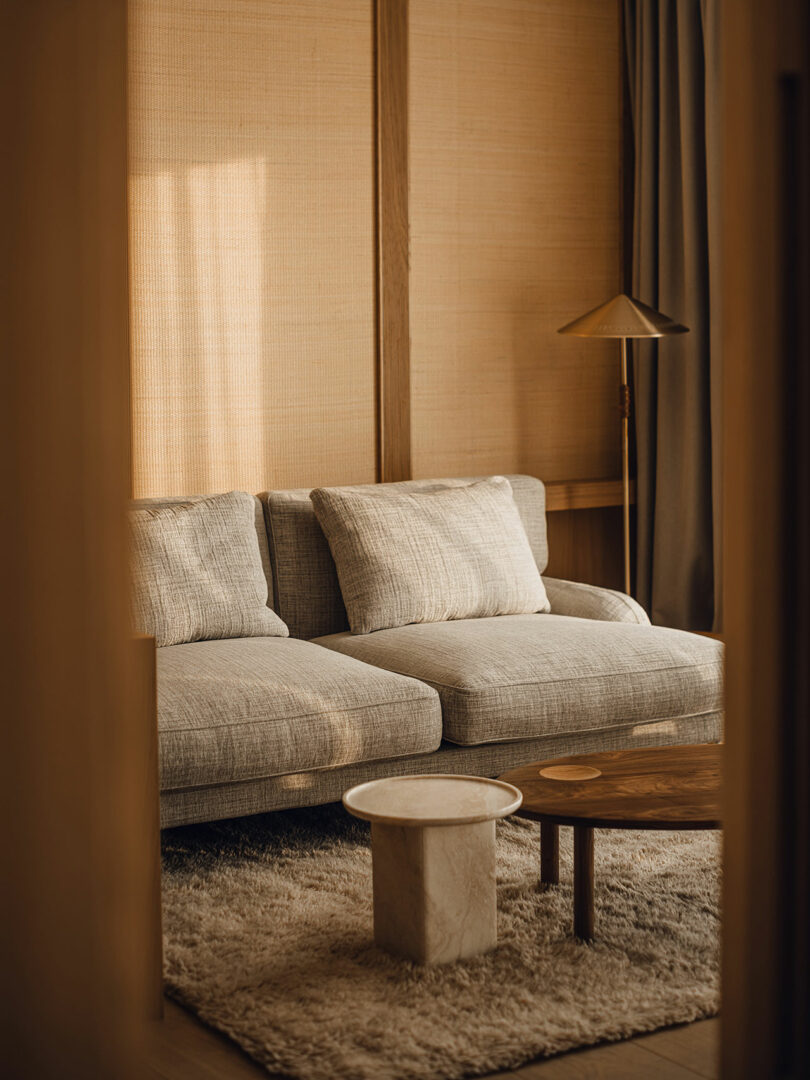 A neutral-toned living room with a gray sofa, a round wooden coffee table, a small marble side table, a floor lamp, and a textured rug.