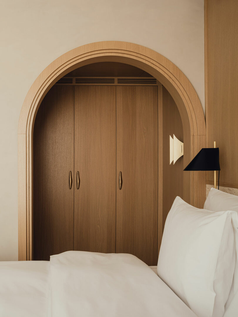 A wooden wardrobe with arched doorway sits behind a neatly made bed with white linens and a black wall-mounted lamp.