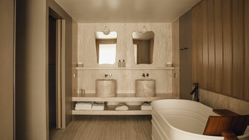 Modern bathroom with two stone sinks, two mirrors, wooden paneling, a white freestanding bathtub, and shelves holding towels and toiletries.