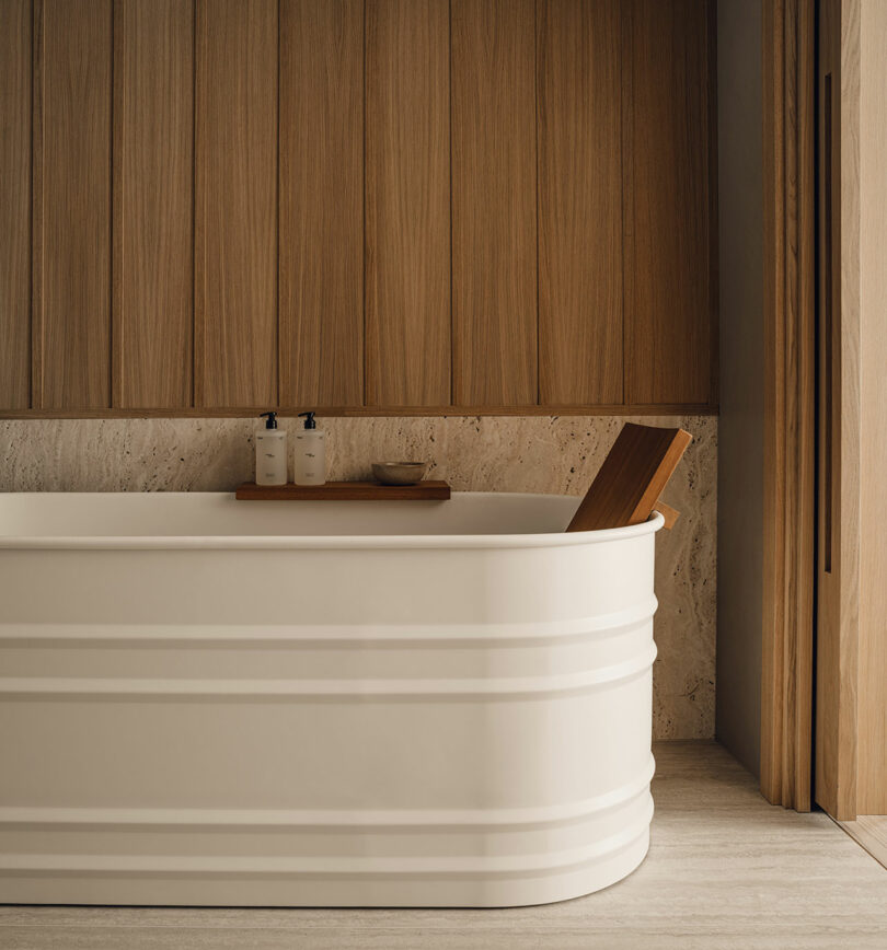 A modern white bathtub with a wooden tray holding two bottles and a bowl, set against wood-paneled walls and stone accents in a minimalist bathroom.