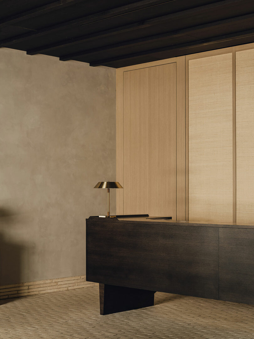 Minimalist office space with a dark wooden desk, brass desk lamp, beige textured walls, and wooden panel accents.