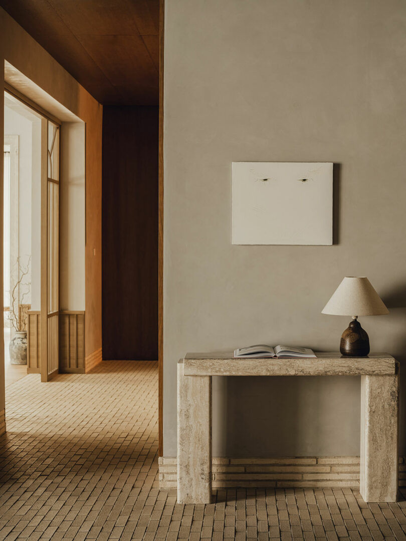 A minimalist hallway with a stone console table, an open book, a ceramic lamp, and a blank white artwork on a beige wall, featuring neutral-toned tile flooring and warm lighting.