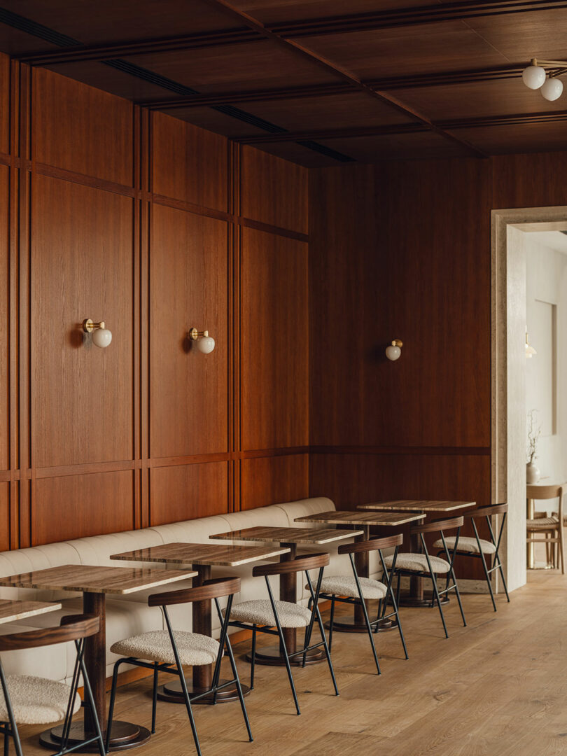 A modern cafe interior with wooden walls, a row of tables and chairs, a beige upholstered bench, and round wall lights.
