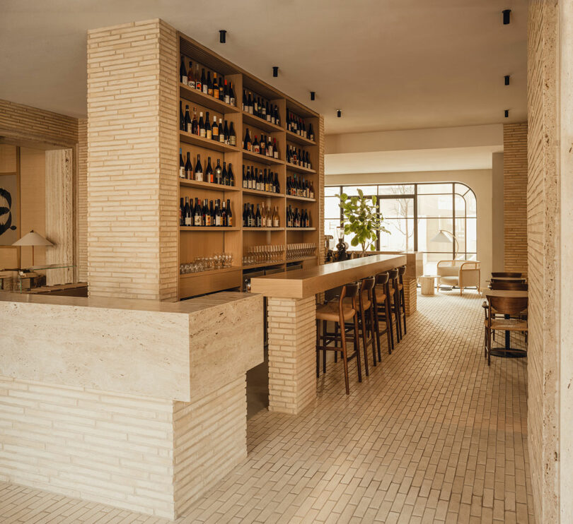 Modern bar interior with light brick walls and floor, a long wooden counter with bar stools, a large wine shelf, and a window letting in natural light.