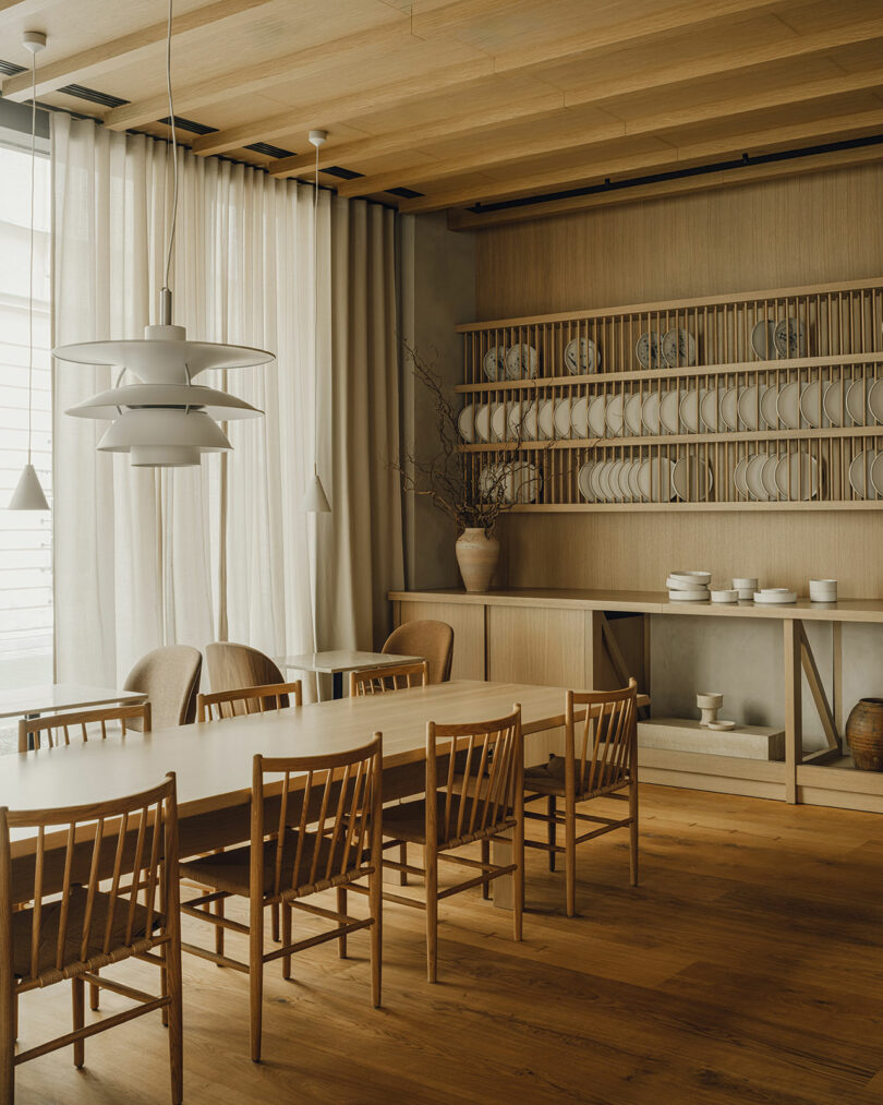 Minimalist dining room with a long wooden table, eight chairs, neutral tones, pendant lights, and open shelves displaying plates and ceramics. Natural light filters through sheer curtains.