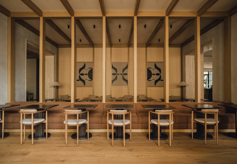 Modern restaurant interior with wooden beams, brown seating, black tables, white chairs, and abstract art on the wall, featuring floor-to-ceiling mirrors dividing the space.