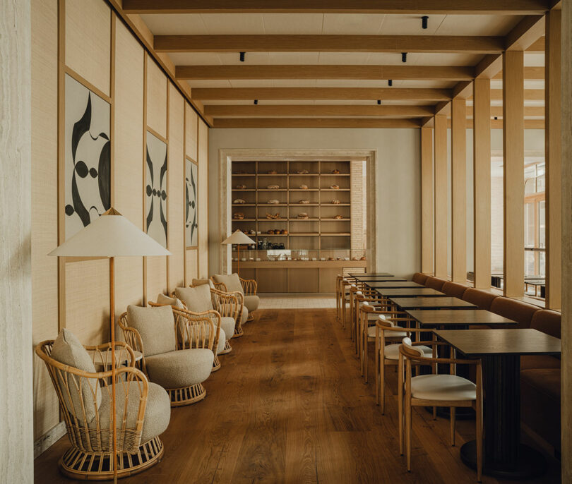 Modern, minimalist lounge with wooden floors, rattan chairs, beige sofas, black tables, and abstract art on the walls; open shelving with pottery at the back.