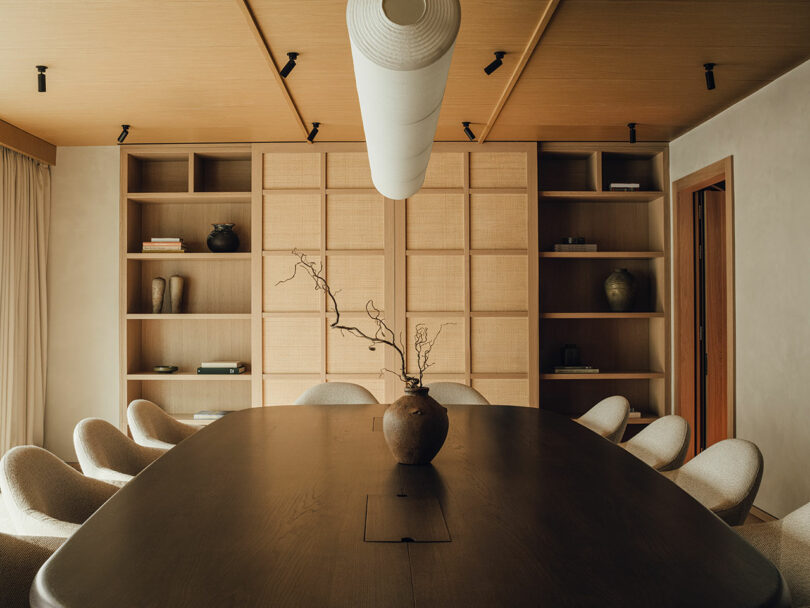 A modern conference room features a large dark wood table, beige upholstered chairs, built-in wooden shelves, and a minimalist vase with branches as a centerpiece.