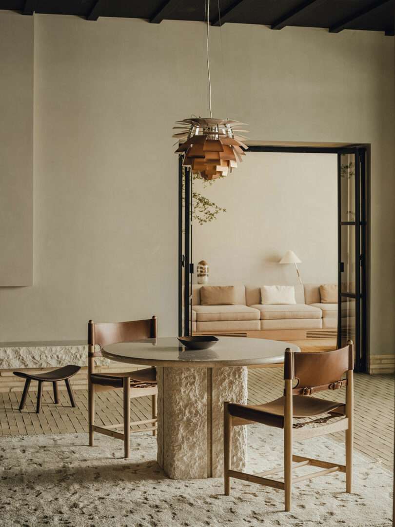 A minimalist dining area with a round stone table, two wooden chairs with brown leather seats, and a modern pendant light, opening to a neutral-toned living room with a sofa and lamp.