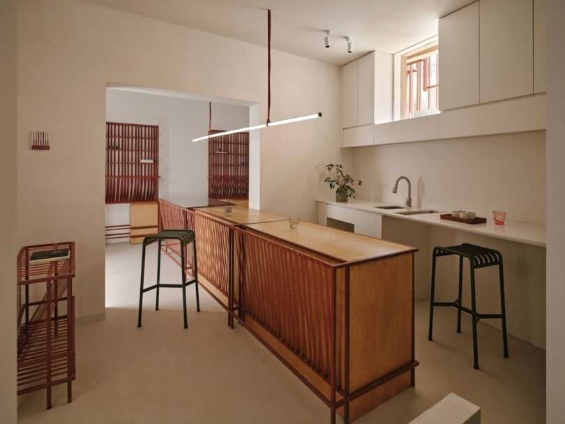 Minimalist kitchen with light wood cabinetry, white countertops, two green bar stools, and a linear ceiling light. Red wood accents and a potted plant are visible. Natural light enters through a small window.