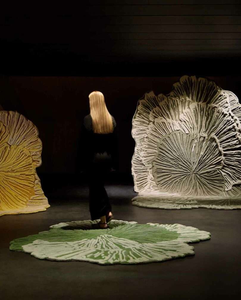 A person with long blonde hair stands on a green, leaf-shaped rug, facing large, textured art pieces resembling leaves in a dimly lit room.