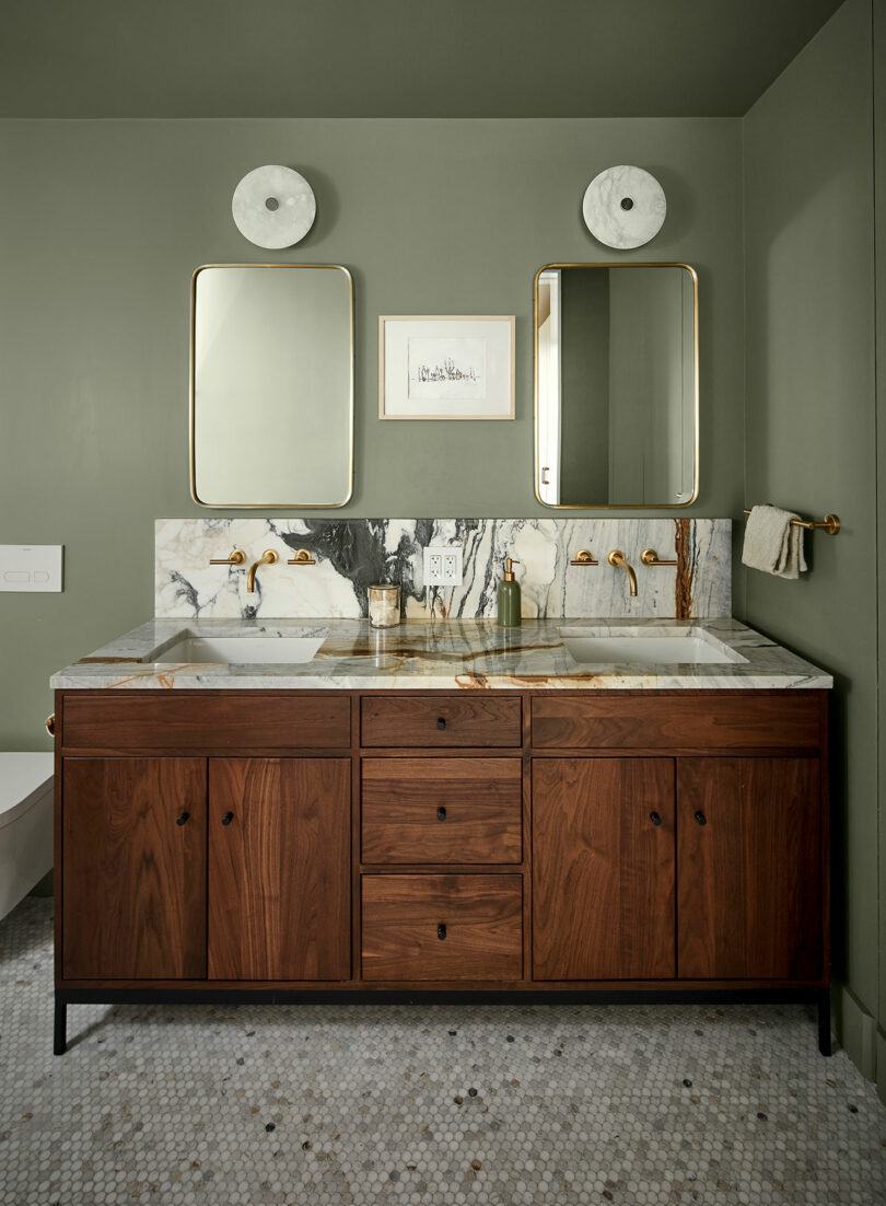 A modern bathroom with a wooden vanity, marble countertop and backsplash, two rectangular mirrors, gold fixtures, and green walls.