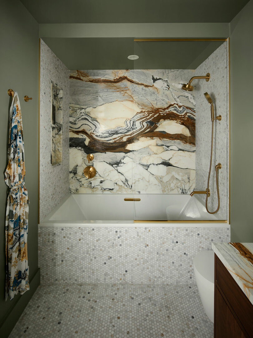 Modern bathroom with a marble tub and shower, gold fixtures, green walls, mosaic tile floor, and a robe hanging on the wall.