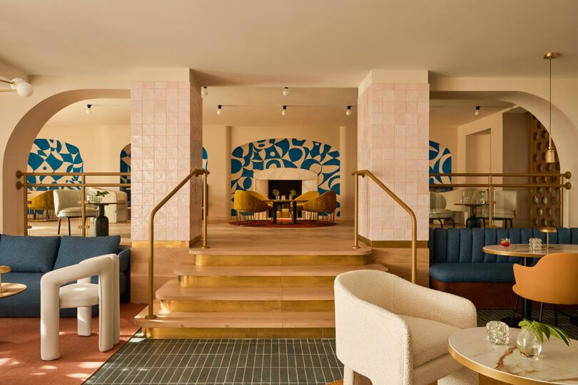 A modern lounge at Kimpton Los Monteros features tiered seating, abstract blue wall art, pastel tiles, gold railings, and a mix of upholstered chairs and sofas arranged around tables.