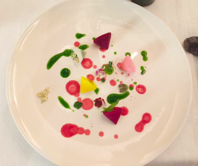 A white plate with colorful geometric pieces of food, green and red sauce dots, and small herb garnishes, arranged artistically on a white tablecloth in the style of Renaud Defrancesco.