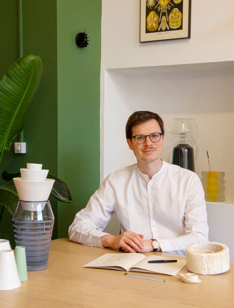 Renaud Defrancesco, a man in glasses and a white shirt, sits at a desk with notebooks, ceramic items, and decorative objects in a modern, green-accented room.