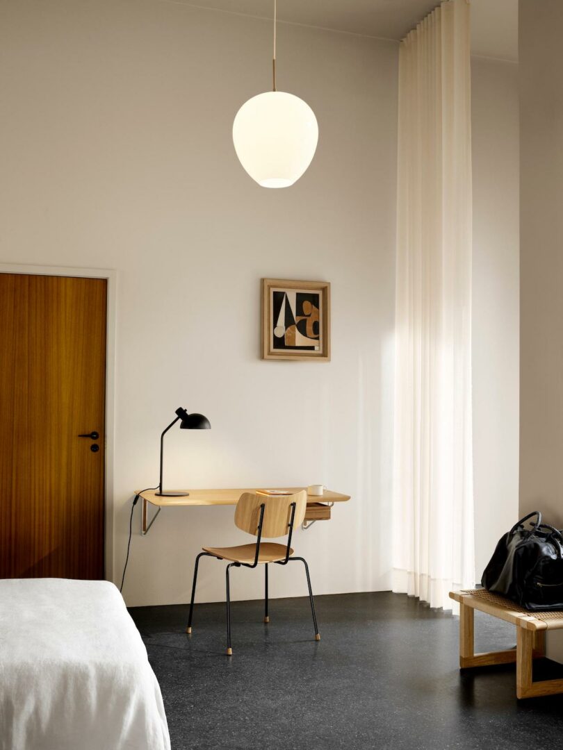A minimalist workspace with a floating wooden desk, Carl Hansen & Son Anker Bak chair, black desk lamp, abstract artwork, and tall white curtains in a bright room