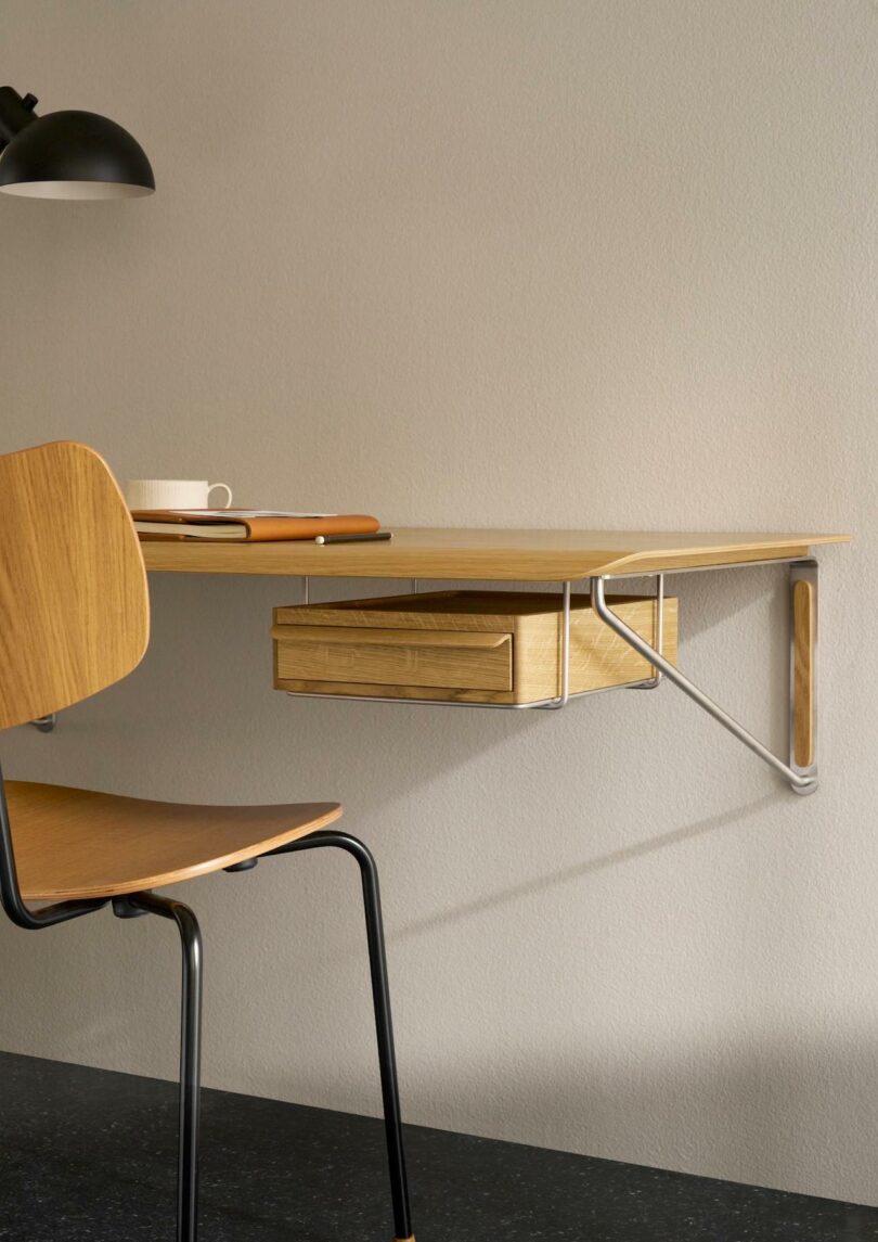 A wall-mounted wooden desk with metal supports, inspired by Carl Hansen & Son Anker Bak, features a drawer, a matching chair, a cup, a notebook, and a black lamp set against a light-colored wall
