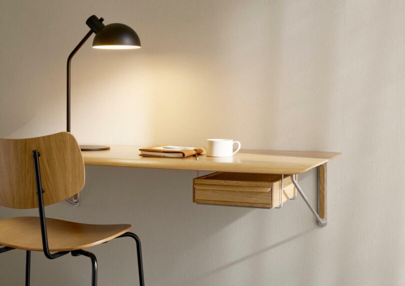 A Carl Hansen & Son Anker Bak wooden chair sits at a minimalist wall-mounted desk with a notebook, pen, white mug, and black desk lamp against a plain beige wall